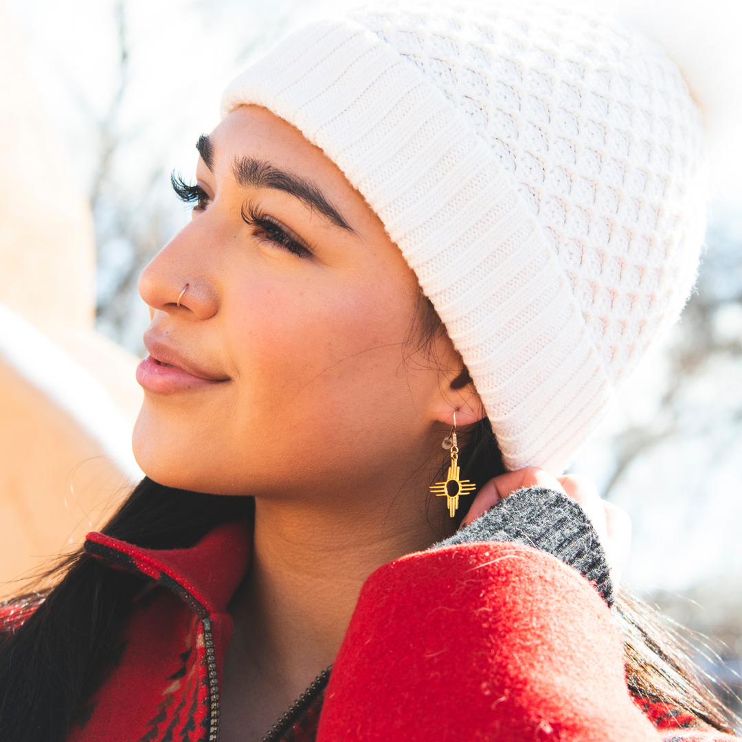Enchantment: Zia Dangle Earrings in sterling silver, featuring a Zia sun symbol, worn by a woman in a white beanie.