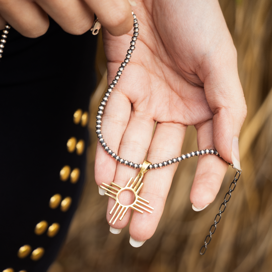 Enchantment: Zia Pendant, sterling silver and gold, held in a hand with a beaded chain, showcasing Native American-inspired design.