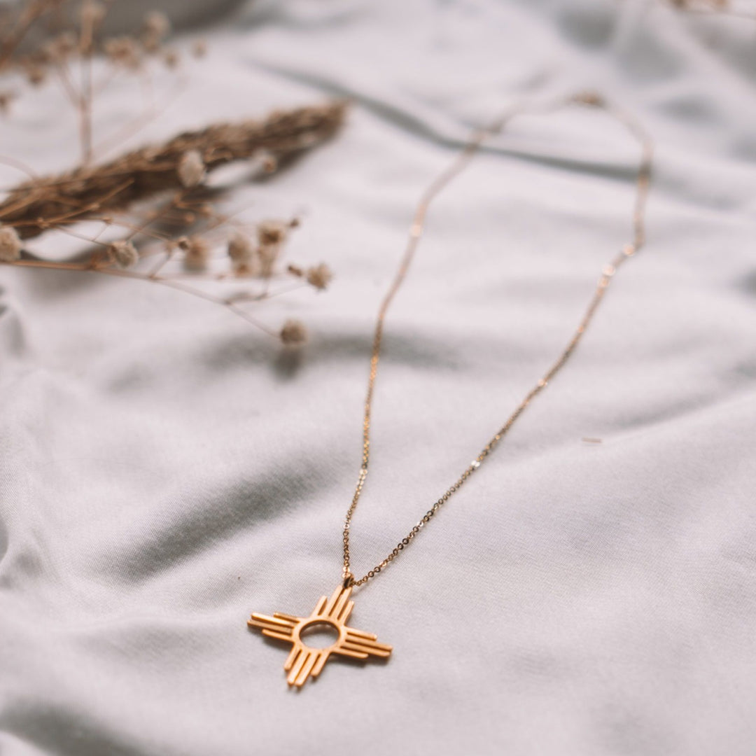 Enchantment: Zia Dangle Necklace in sterling silver, featuring a Zia symbol on a delicate chain, resting on soft fabric.