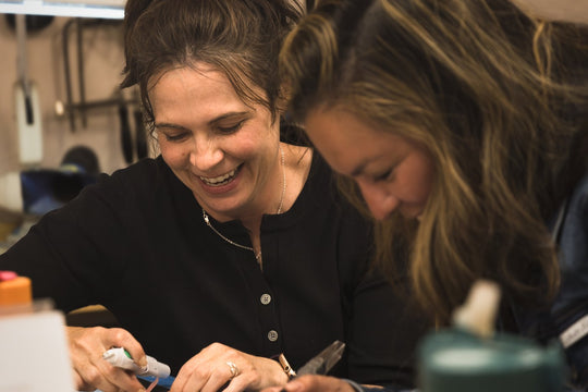 Two Craft & Cast participants laughing together as they carve their wax jewelry pieces at the TSkies workshop bench.
