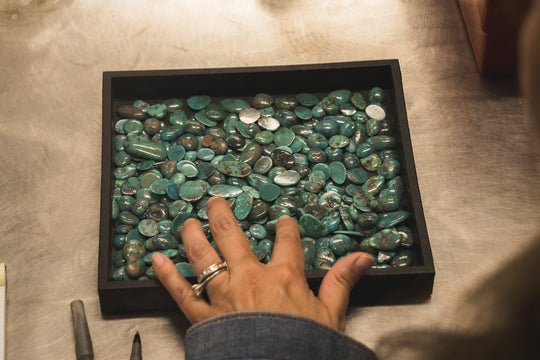 Hand reaching into a tray of polished turquoise stones at the TSkies workshop bench.